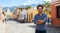 Young Hispanic male in striped shirt standing confidently on cobblestone street with colorful colonial architecture and mountains in background. Travel and lifestyle portrait.