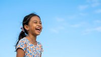 Young girl with dark hair and South Asian ethnicity wearing a blue floral print shirt, smiling outdoors against a clear blue sky. Suitable for family, lifestyle, and children's content.