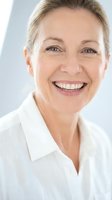 Mature Caucasian woman with blonde hair wearing white collared shirt, smiling warmly at camera against neutral background. Corporate portrait for business, healthcare, or professional services.