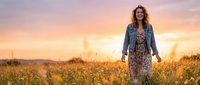 Middle-aged woman with long wavy brown hair and fair complexion wearing denim jacket and floral dress standing in golden wheat field at sunset for lifestyle and nature photography.