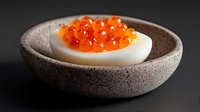 Gourmet hard-boiled egg topped with bright orange caviar served in rustic ceramic bowl on dark background for luxury food photography and culinary content.
