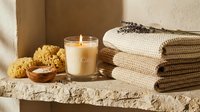 Lit candle with sea sponge, bath salts, lavender, and stacked waffle towels on stone shelf in warm spa setting.