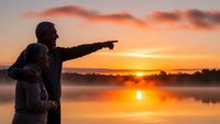 Father and son silhouettes pointing at golden sunrise over calm lake water with dramatic orange sky clouds reflecting on surface during peaceful morning.