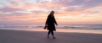 Young woman in dark winter coat walking alone on sandy beach at sunset, suitable for lifestyle, travel, and wellness editorial use.