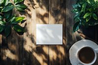 Blank white paper card with natural sunlight shadows, coffee cup and green leaves on rustic wooden table. Morning creative workspace layout.