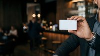 Businessman holding blank white business card in modern bar or restaurant setting with blurred background and warm lighting for corporate branding and networking purposes.