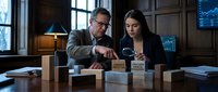 Middle-aged Caucasian businessman and young Hispanic businesswoman analyzing wooden blocks in modern office setting for strategic planning and teamwork.