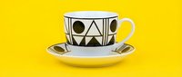 Geometric patterned teacup and saucer with black triangles and circles on white ceramic, yellow handle and bright yellow background, modern tableware design.