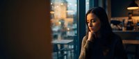 Young Asian woman in cozy sweater looking thoughtfully out rainy cafe window at night with warm ambient lighting creating peaceful contemplative mood.