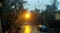 Raindrops on window glass with blurred street lights creating golden bokeh effect during evening storm, moody atmospheric scene.