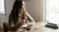 Young woman enjoying healthy breakfast bowl with berries at wooden table by window in cozy morning light for wellness lifestyle content.