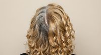 Blonde woman with curly hair showing gray roots needing touch-up, back view against neutral background for hair care and beauty salon concepts.