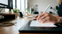 Designer using stylus pen on digital tablet at wooden desk with plants and windows in modern office workspace for creative work and design projects.