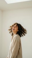 Happy young woman with curly hair laughing and moving in beige linen shirt against neutral wall background expressing joy and freedom in minimalist interior.