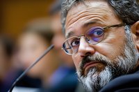 Middle-aged professional man with salt and pepper beard wearing purple glasses during conference or hearing, expressing thoughtful concern against blurred background.