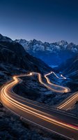 Winding mountain road with golden light trails from vehicle headlights and taillights at dusk, snow-covered alpine peaks in background, long exposure night photography for travel and transportation concepts.