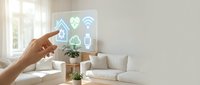 Hand touching transparent smart home control panel displaying connected device icons in modern living room with white furniture and natural light from window.