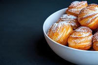 Fresh homemade profiteroles dusted with powdered sugar served in white ceramic bowl against dark background, close up view of French pastries.