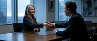 Professional businesswoman and businessman shaking hands across conference table in modern office meeting room with city skyline view through windows.
