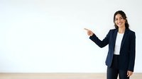 Young woman with dark hair and South Asian ethnicity wearing navy blazer and white shirt, pointing to blank white wall space with smile, suitable for business presentations, advertising copy, and corporate marketing materials.