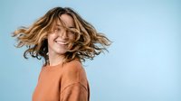 Happy young Caucasian woman with flowing blonde hair wearing orange sweater laughing with joy against light blue background for lifestyle content.