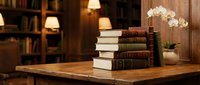 Stacked vintage books with white orchid flowers on wooden desk in warm library setting with bookshelves and lamp.
