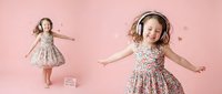 Young girl with blonde hair dancing joyfully in floral dress against pink background. Child wearing headphones expressing happiness and freedom through movement.