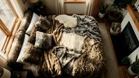 Cozy unmade bed with rumpled beige linens and pillows in sunlit bedroom with wooden frames and plants creating warm morning atmosphere.