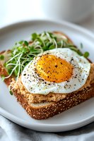 Sunny side up egg on sesame seeded bread with microgreens and black pepper, served on white plate. Fresh breakfast sandwich with crispy edges and runny yolk.