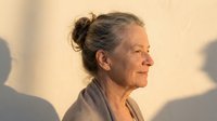 Mature woman with gray hair in bun, profile view, wearing neutral beige clothing against warm background with soft shadows, suitable for aging, wellness, and lifestyle editorial use.