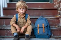 Young boy with blonde hair sitting on brick steps wearing beige outfit with blue backpack beside him, ready for school or adventure.