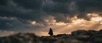 Silhouette of person walking on rocky terrain against dramatic stormy sky with golden sunlight breaking through dark clouds creating moody atmospheric scene.