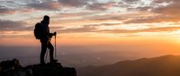 Silhouette of hiker with backpack and trekking poles standing on mountain peak at golden sunset, overlooking vast valley landscape below.