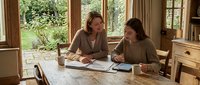 Two professional women collaborating on business documents at wooden table in bright home office with garden view through large windows.