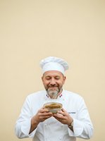 Professional male chef with gray beard wearing white chef's coat and hat holding bowl of soup against neutral beige background. Culinary professional displaying prepared dish for restaurant, cooking school or food service marketing.
