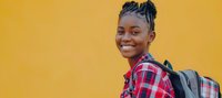Young African American woman with braided hairstyle wearing red plaid shirt and backpack smiling confidently against vibrant yellow wall background, expressing joy and optimism.