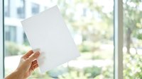 Cropped hand holding blank white paper sheet against bright window with urban cityscape and green foliage visible in background, suitable for mockup and advertising.
