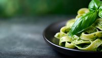 Fresh fettuccine pasta with basil pesto sauce served in dark ceramic bowl on black stone background with copy space, selective focus and green bokeh.