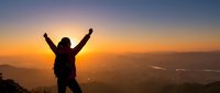 Silhouette of victorious woman hiker with raised arms celebrating success on mountain summit at golden sunrise with panoramic valley view below.