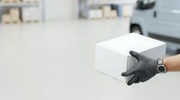 Delivery worker wearing black gloves holding white cardboard box in warehouse or logistics facility with vehicles in background.