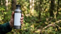 Unisex hand holding reusable water bottle with blank label in forest setting, promoting eco-friendly hydration and sustainable lifestyle choices outdoors.
