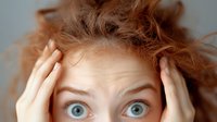 Young woman with bright blue eyes and messy ginger hair holding head in hands with shocked surprised expression against light background.