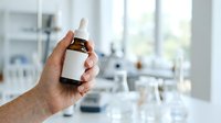 Male hand holding brown dropper bottle with white label in modern laboratory or medical facility setting for skincare, wellness, or pharmaceutical product presentation.