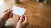Hands holding blank white card on wooden table surface, suitable for business card mockups, branding presentations, and corporate identity designs.