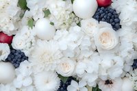Lush floral arrangement with white roses, dahlias, hydrangeas, blueberries, red apples, and white spheres for wedding or event decor.