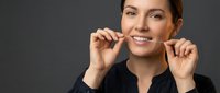 Woman with brown hair and fair complexion flossing teeth against neutral gray background, promoting dental hygiene and oral care routines for health and wellness content.
