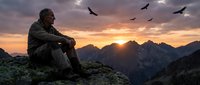 Contemplative hiker sits on mountain peak watching sunset over alpine landscape with soaring birds overhead during golden hour adventure.