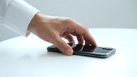 Male hand in white shirt sleeve reaching for a black smartphone lying on a white surface, close-up side view.