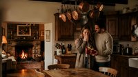 Young Caucasian couple cooking together in rustic kitchen with fireplace, sharing intimate moment while preparing meal in cozy home interior.