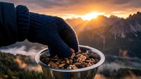 Hiker in knitted glove pouring trail mix into metal bowl with mountain sunrise and misty valley backdrop, outdoor adventure and camping food preparation.